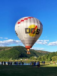 Hei&szlig;luftballon beim Start am Badesee mit Zuschauern vor der Kulisse des bewaldeten Honigberg.